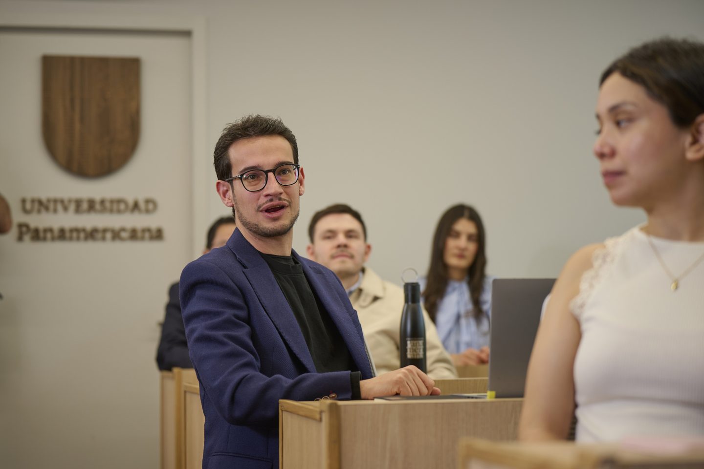 Profesor conversa con dos estudiantes frente al escudo de la Universidad Panamericana